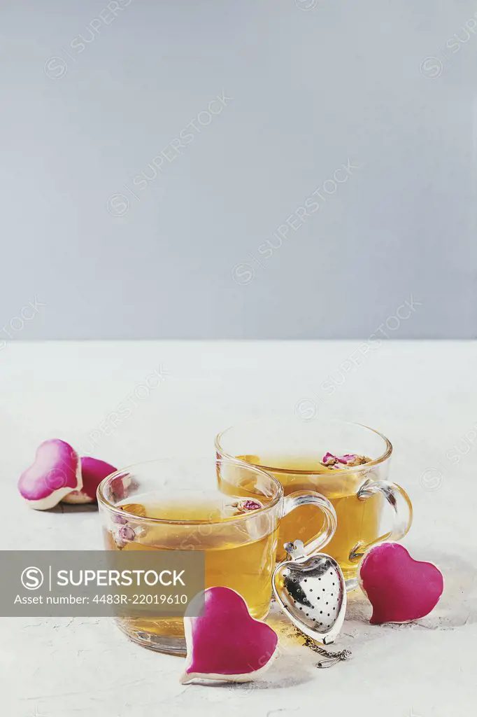 Love Valentines day greeting card with two glasses of hot tea and heart shape homemade cookies with pink icing and tea strainer over white table. Copy space. Toned image