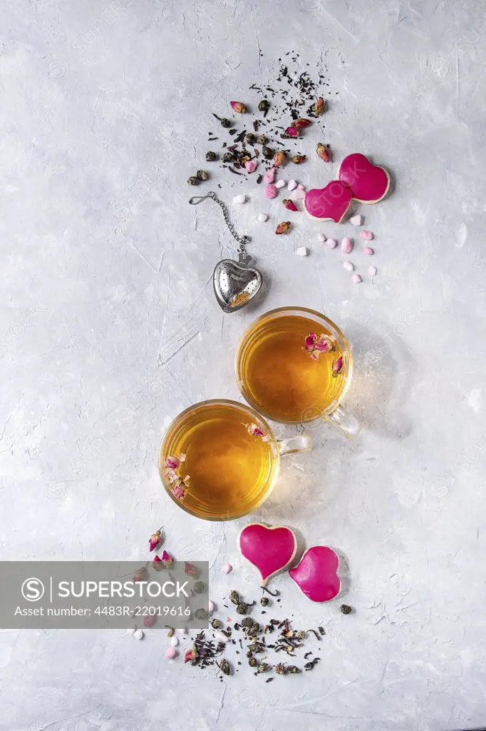 Love Valentines day greeting card with two glasses of hot tea, rose buds, heart shape homemade cookies as gift, pink sugar, tea strainer over gray texture background. Top view, space