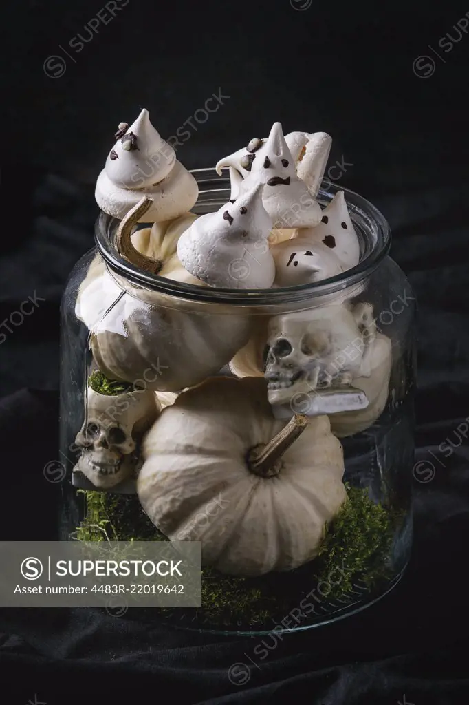 Halloween sweet dessert table decor glass jar with white meringue ghosts with chocolate eyes, decor skulls, moss and pumpkin over black background.