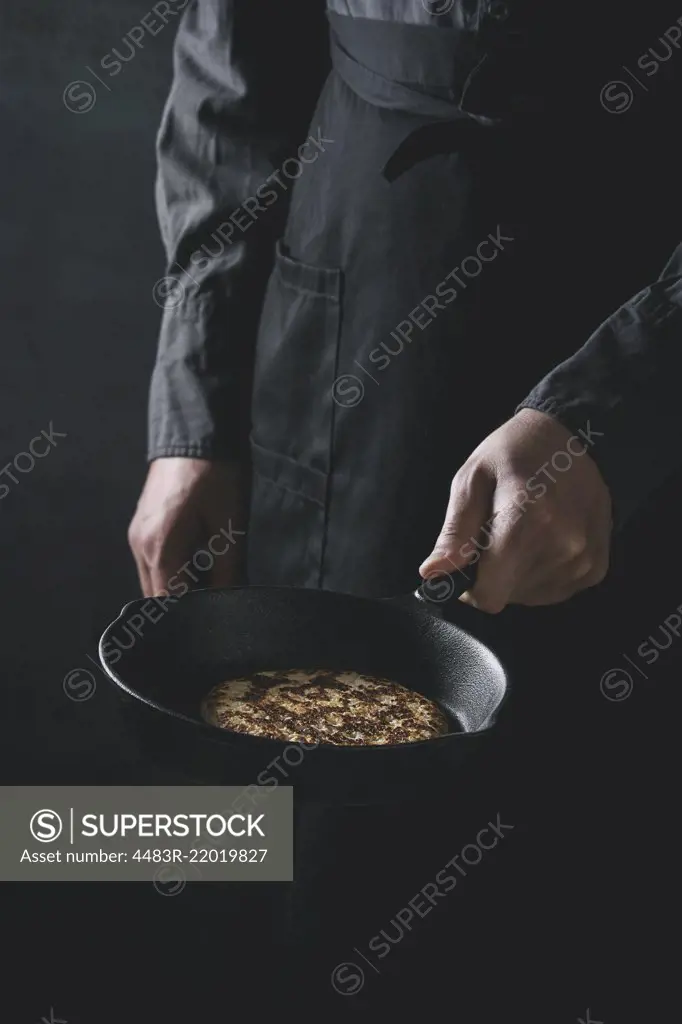 Man chef in black apron cooking pancakes in cast-iron pan. Holding pan in hand. Dark rustic style. Toned image