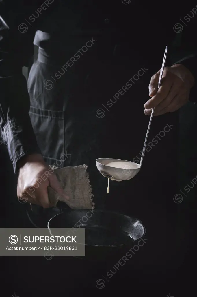 Man chef in black apron pouring dough from ladle for cooking pancakes in cast-iron pan. Dark rustic style.