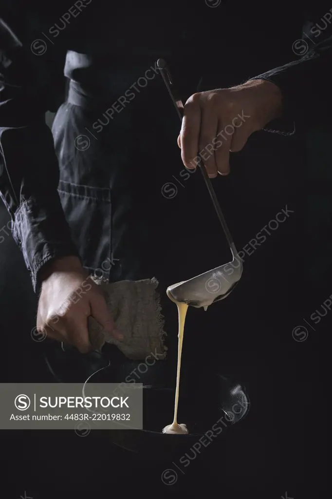 Man chef in black apron pouring dough from ladle for cooking pancakes in cast-iron pan. Dark rustic style.