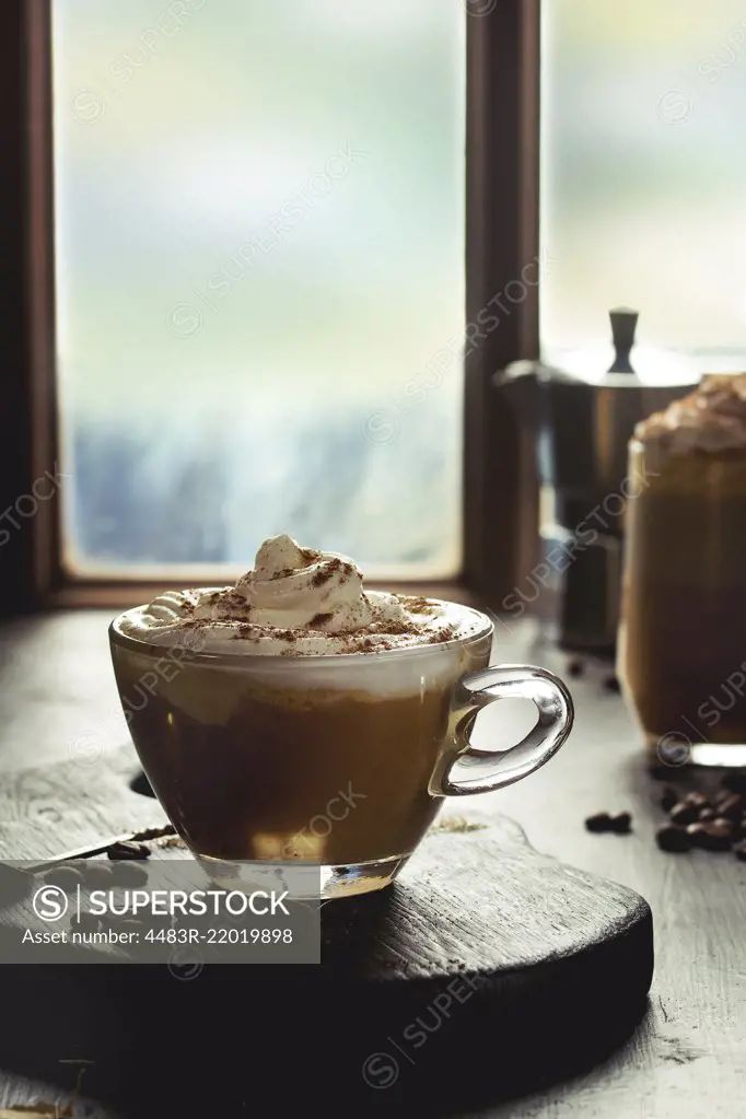 Glass of spicy pumpkin latte with whipped cream and cinnamon standing wooden serving board on cafe table near window with coffeepot and coffee beans above. Atmospheric breakfast