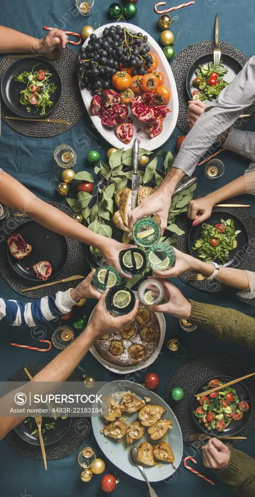 Company of friends gathering for Christmas or New Year party dinner at festive table. Flat-lay of hands holding glasses with drinks, feasting and celebrating holiday, top view, vertical composition