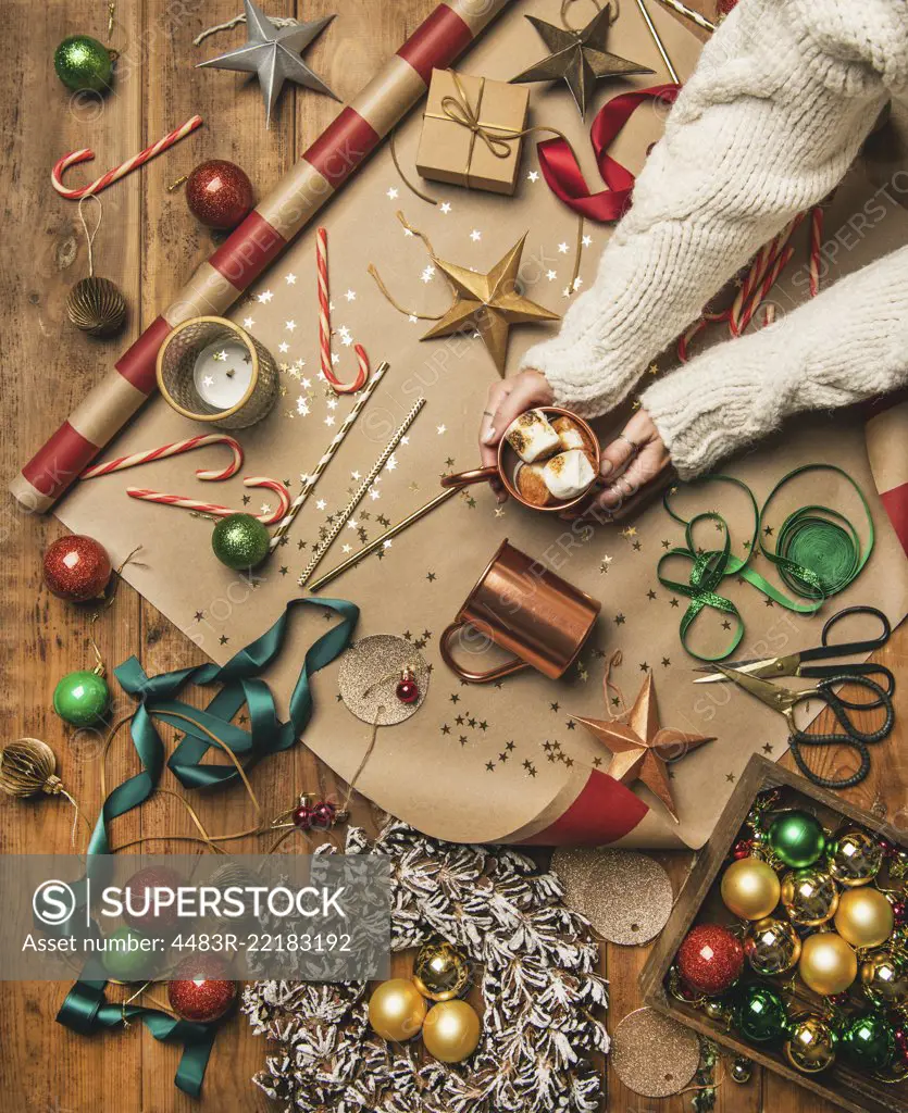 Flat-lay of hot chocolate drink with toasted marshmallows in woman's hands, Christmas decorations, door wreath, ribbons, gift paper, glittering balls, candy canes on wooden background, top view