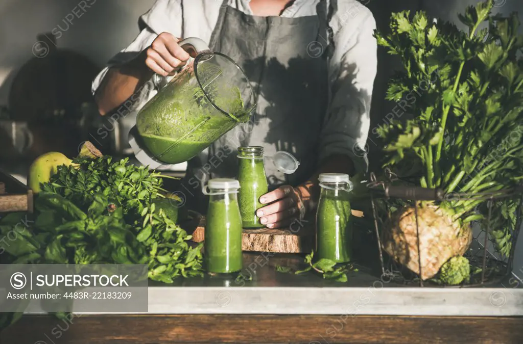 Making green detox take-away smoothie. Woman in linen apron pouring green smoothie drink from blender to bottle surrounded with vegetables and greens. Healthy, clean eating, weight loss food concept