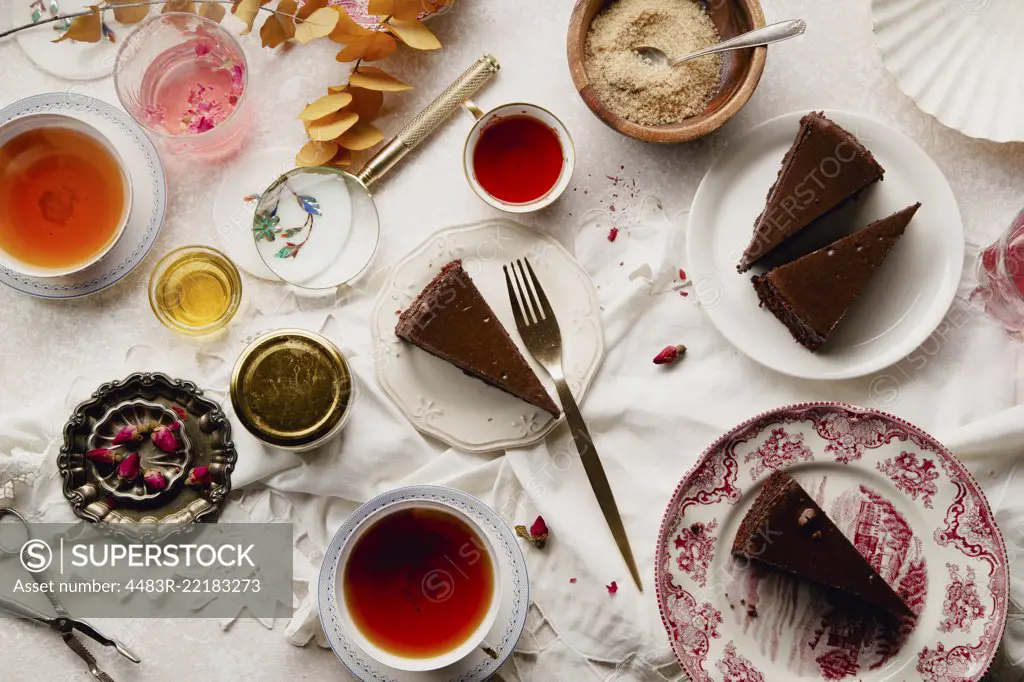 Overhead image of tea party with chocolate cheesecake slices on various plates. Traditional style, vintage tablecloth and cutlery.