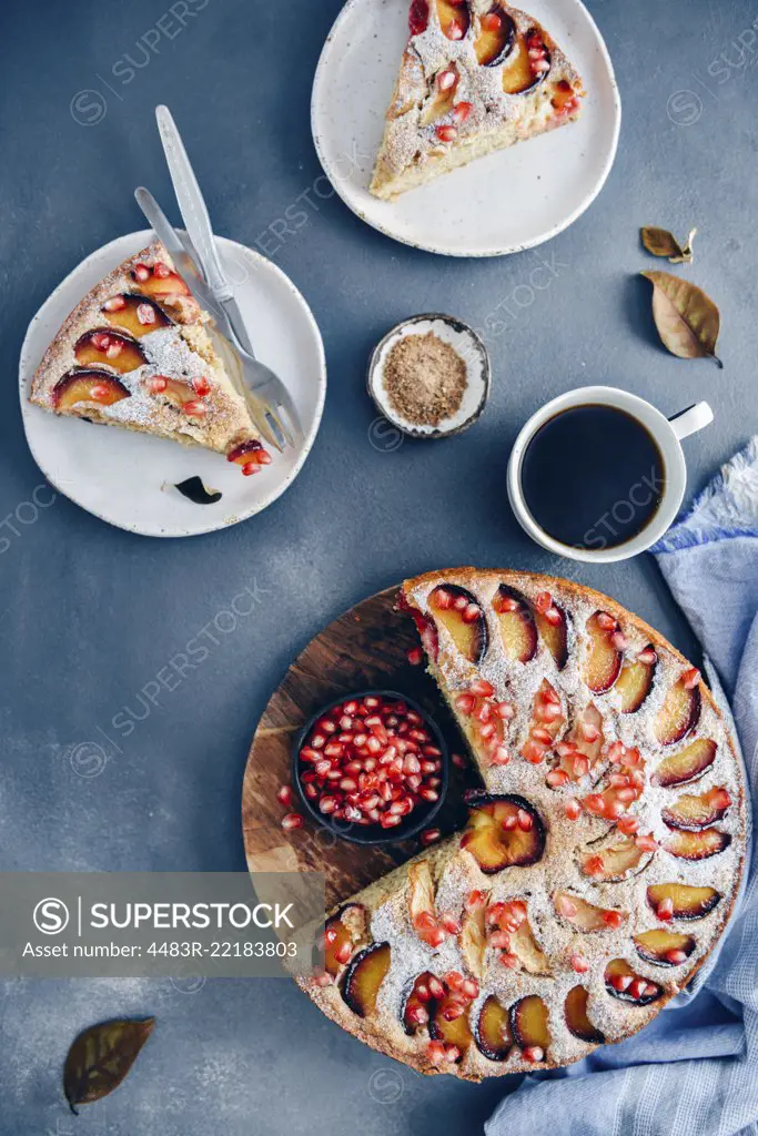 Apple plum cake topped with slices of apple and plum photographed on a dark backdrop. Slices of cake on two white plates, a cup of coffee and a small of brown sugar accompany.