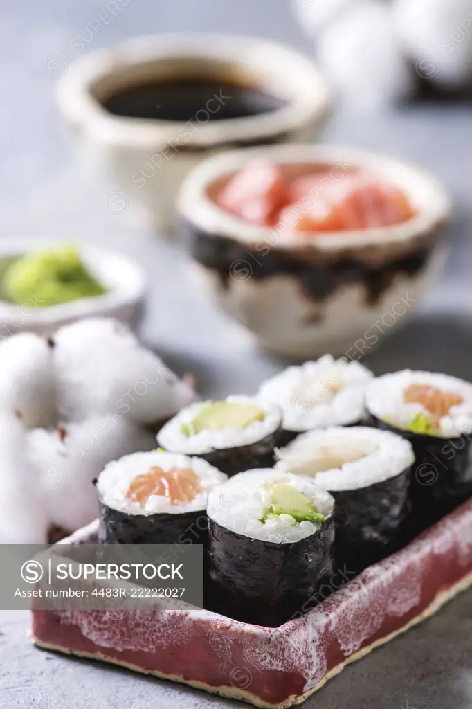 Sushi rolls set in pink ceramic serving plate with bowls of soy sauce and pickled ginger, cotton flowers on light blue table. Japan menu. Close up