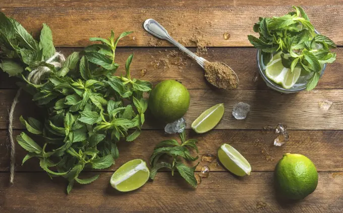 Ingredients for making mojito summer cocktail. Fresh mint bunch, limes, brown sugar and ice over rustic wooden background, top view