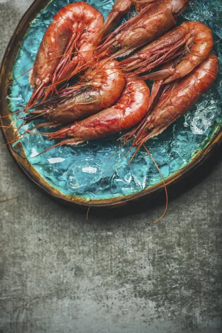 Raw uncooked red shrimps on chipped ice in turquoise blue ceramic tray over grey concrete background, top view, copy space, vertical composition. Fresh seafood concept