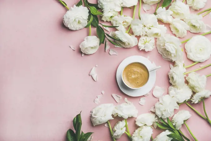 Spring morning concept. Flat-lay of cup of coffee surrounded with white ranunculus flowers over light pink background, top view, copy space, horizontal composition