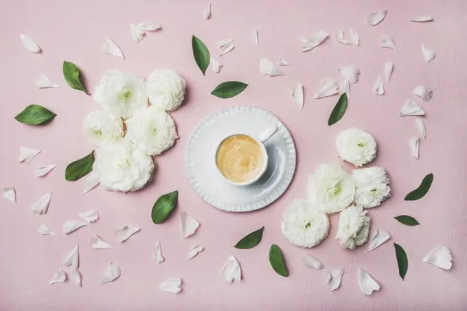 Spring morning concept. Flat-lay of cup of coffee surrounded with white ranunculus flowers and petals over light pink pastel background, top view