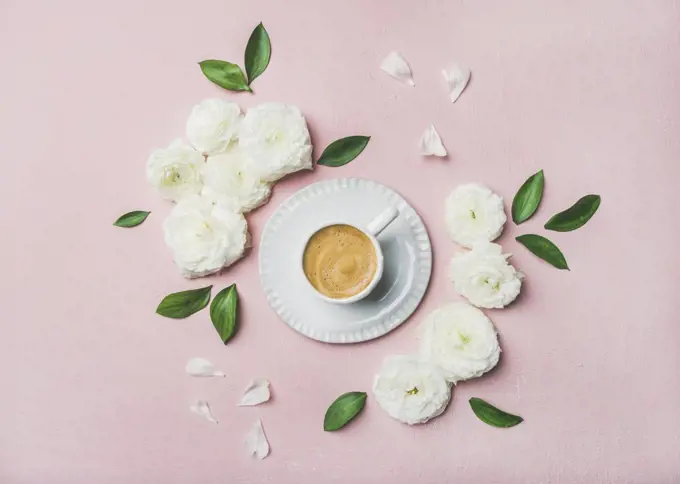 Spring morning concept. Flat-lay of cup of freshly brewed coffee surrounded with white ranunculus flowers and petals over light pink pastel background, top view