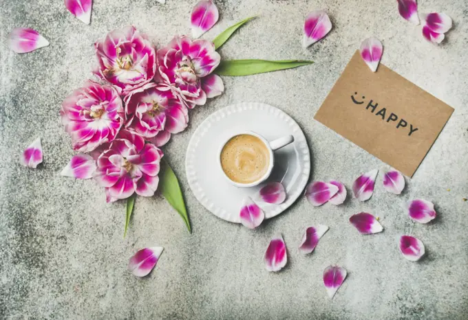 Spring morning concept. Flat-lay of cup of coffee surrounded with pink tulip flowers and sign happy on paper over grey marble background, top view