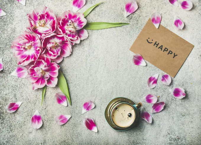 Spring morning concept. Flat-lay of cup of coffee surrounded with pink tulip flowers, petals and sign happy on paper over grey marble background, top view, copy space