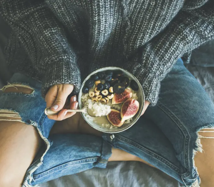 Healthy winter breakfast in bed. Woman in woolen sweater and shabby jeans eating rice coconut porridge with figs, berries, hazelnuts, top view. Clean eating, vegetarian, comfort food concepts