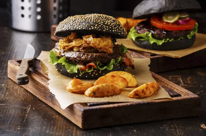 Black burger with sesame seed bun, meat, bacon, onion fries rings and potato wedges on dark wooden background