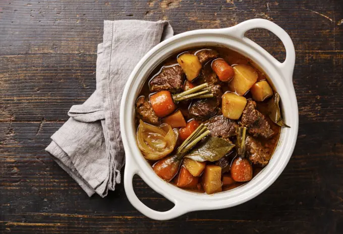 Beef meat stewed with potatoes, carrots and spices in ceramic pot on wooden background