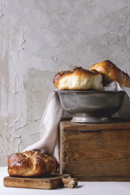 Heap of sweet round sabbath challah bread with white and black sesame seeds in vintage metal bowl on wooden chest and on small cutting board over white table with plastered wall at background.