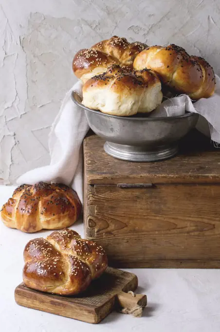 Heap of sweet round sabbath challah bread with white and black sesame seeds in vintage metal bowl on wooden chest and on small cutting board over white table with plastered wall at background.