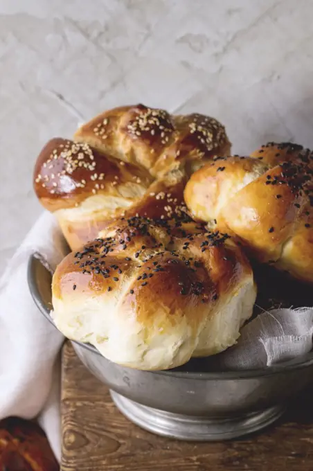 Heap of sweet round sabbath challah bread with white and black sesame seeds in vintage metal bowl on wooden table with plastered wall at background.