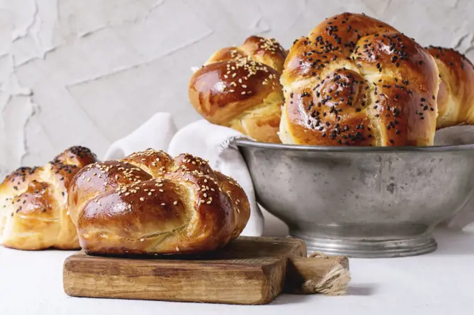 Heap of sweet round sabbath challah bread with white and black sesame seeds in vintage metal bowl and on small cutting board over white table with plastered wall at background.