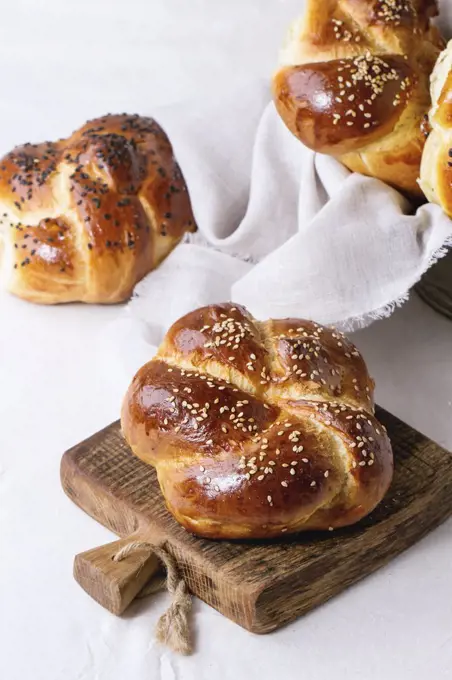 Heap of sweet round sabbath challah bread with white and black sesame seeds on small cutting board over white table with plastered wall at background.