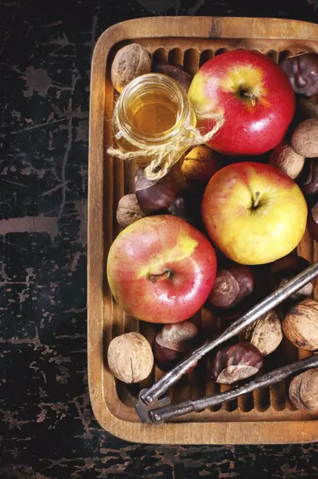 Glass jar of honey with apples, chestnuts and walnuts in wooden plate over black wooden table. Top view