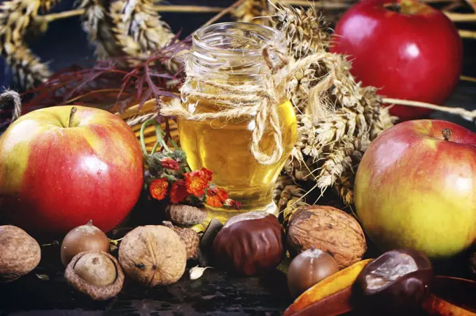 Glass jar of honey with apples, ears of wheat, chestnuts and walnuts over black wooden table