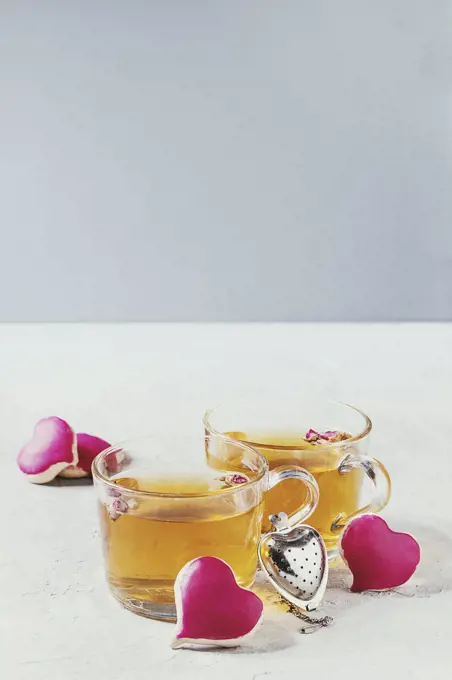 Love Valentines day greeting card with two glasses of hot tea and heart shape homemade cookies with pink icing and tea strainer over white table. Copy space. Toned image