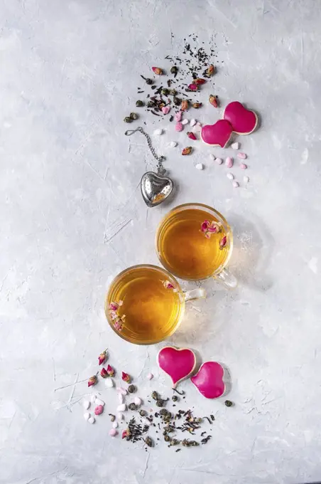 Love Valentines day greeting card with two glasses of hot tea, rose buds, heart shape homemade cookies as gift, pink sugar, tea strainer over gray texture background. Top view, space