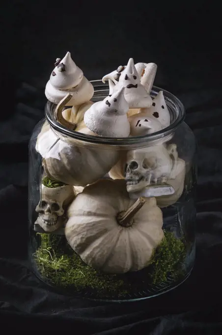 Halloween sweet dessert table decor glass jar with white meringue ghosts with chocolate eyes, decor skulls, moss and pumpkin over black background.