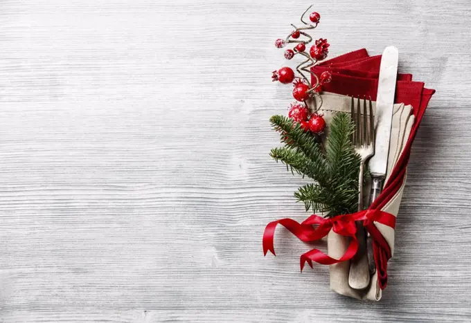 Table fork and knife set with napkin, christmas fir branch, red berries and ribbon on gray wooden background copy space