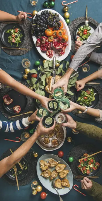 Company of friends gathering for Christmas or New Year party dinner at festive table. Flat-lay of hands holding glasses with drinks, feasting and celebrating holiday, top view, vertical composition