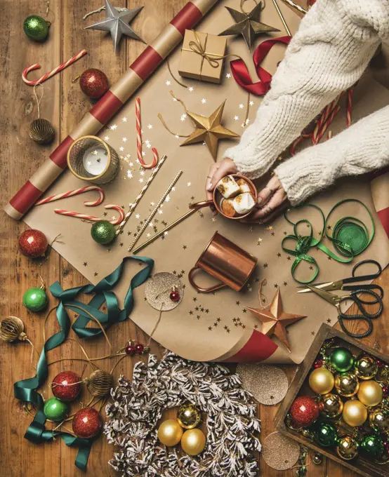 Flat-lay of hot chocolate drink with toasted marshmallows in woman's hands, Christmas decorations, door wreath, ribbons, gift paper, glittering balls, candy canes on wooden background, top view