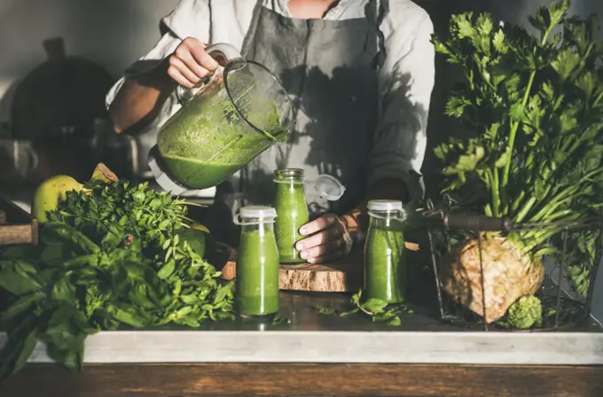 Making green detox take-away smoothie. Woman in linen apron pouring green smoothie drink from blender to bottle surrounded with vegetables and greens. Healthy, clean eating, weight loss food concept