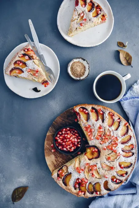 Apple plum cake topped with slices of apple and plum photographed on a dark backdrop. Slices of cake on two white plates, a cup of coffee and a small of brown sugar accompany.