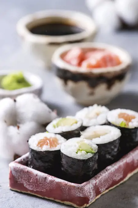 Sushi rolls set in pink ceramic serving plate with bowls of soy sauce and pickled ginger, cotton flowers on light blue table. Japan menu. Close up