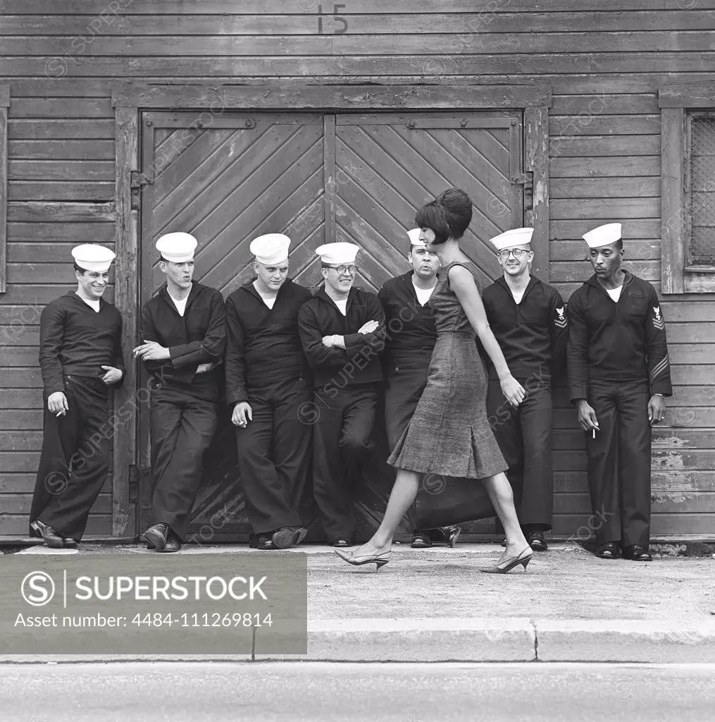 Fashion in the 1960s. A group of American sailors are standing at the docks on shore leave and makes a nice addition to the fashion shoot. A young model passes them wearing a dress with matching handbag and shoes. Sweden 1964 ref BV59-5