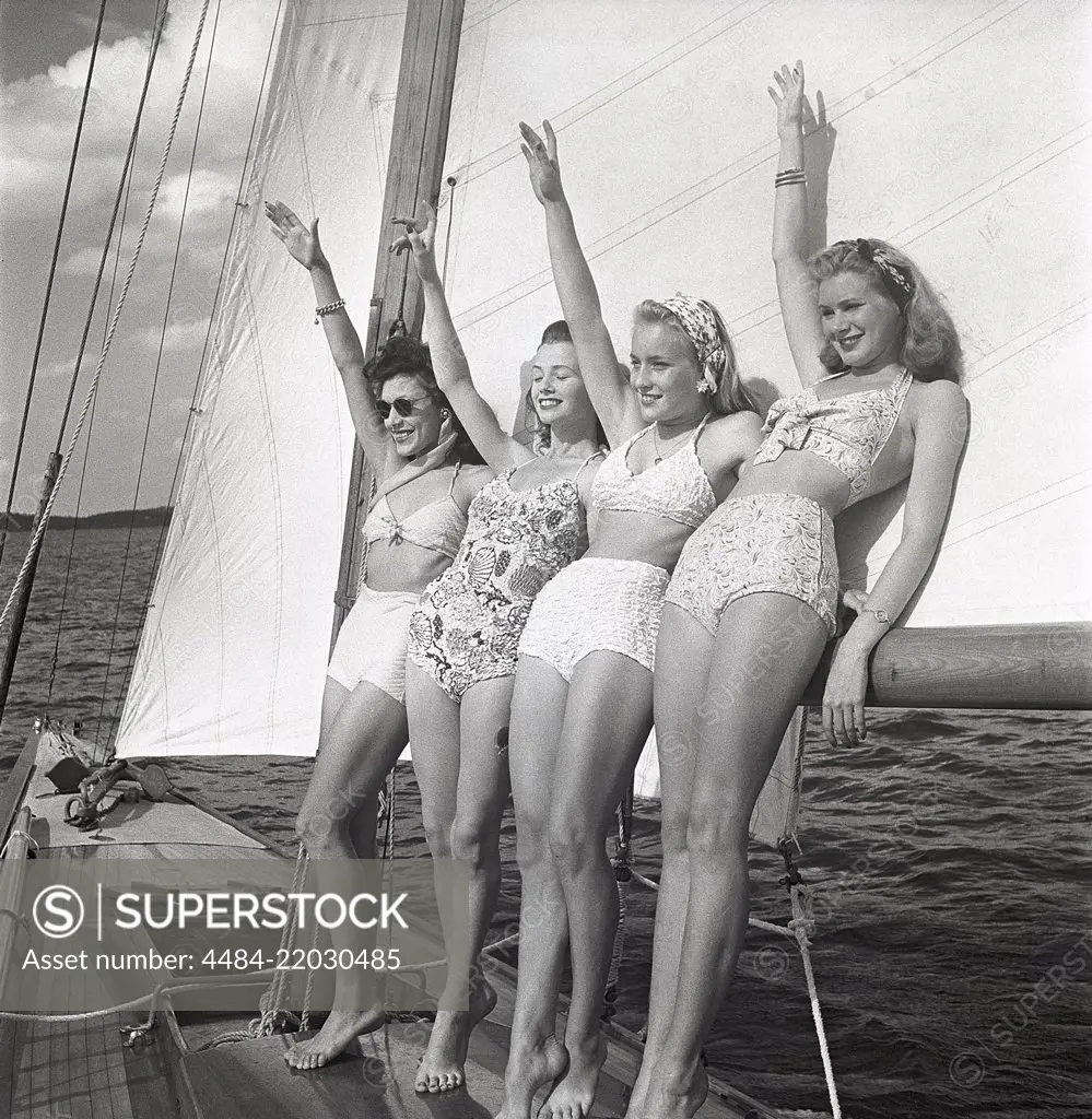 1940s sailing boat. Four young women are standing and leaning against the sails of the boat waiving as you should when you meet another boat. They are wearing the typical 1940s bathingsuit and bikini with high waist. Sweden 1946 Photo Kristoffersson ref AC100-4