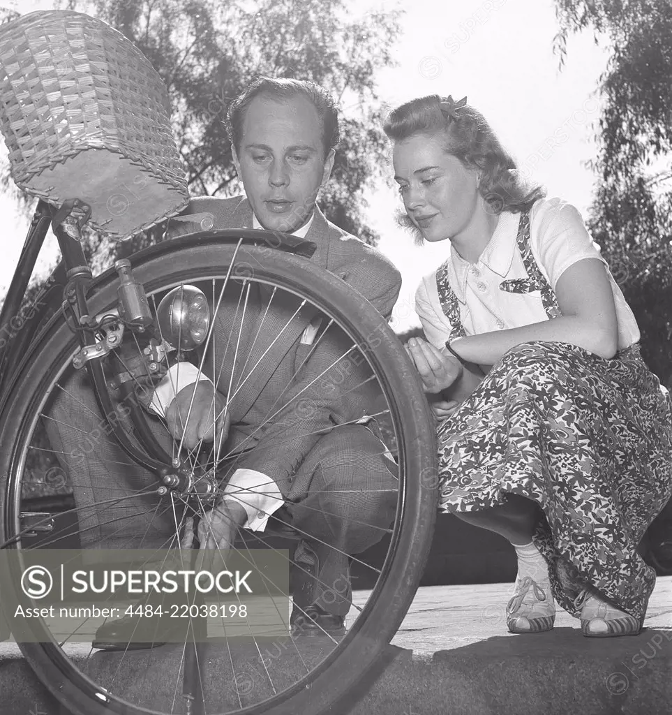 1940s couple with a bicycle. Actor Willy Peters is seen here pumping air into the tire of a womans bicycle while she is sitting beside him and looking. Sweden 1940s. Photo Kristoffersson E50-3