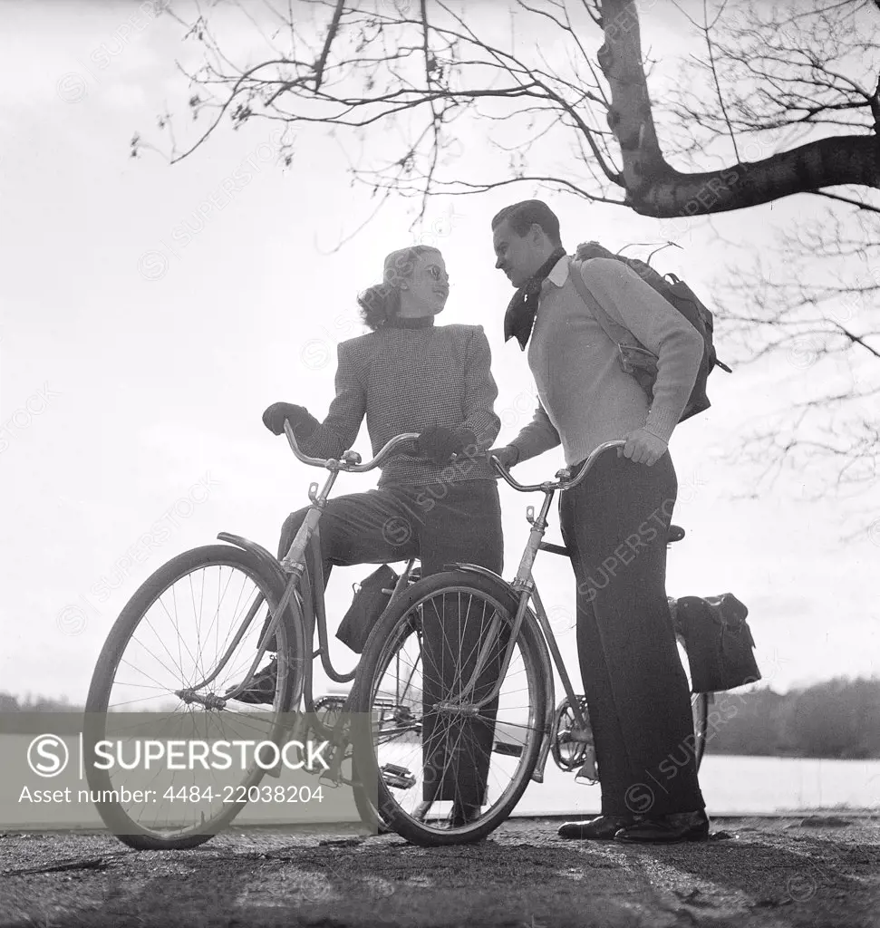 1940s couple on bicycles. A young couple is out on a tour on their bicycles on a sunny spring day. They have practical bags attached to the racks of the bicycles where they could transport the picnic food. Sweden 1947. Photo Kristoffersson AB11-10