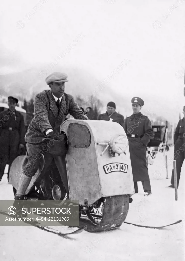 Odd winter vehicle. A man is sitting on his home made snow mobile. A simple construction of a motorcycle inside a wooden body, with a belt instead of wheels to float on the snow surface. The balance and steering is made with the skiis. 1930s