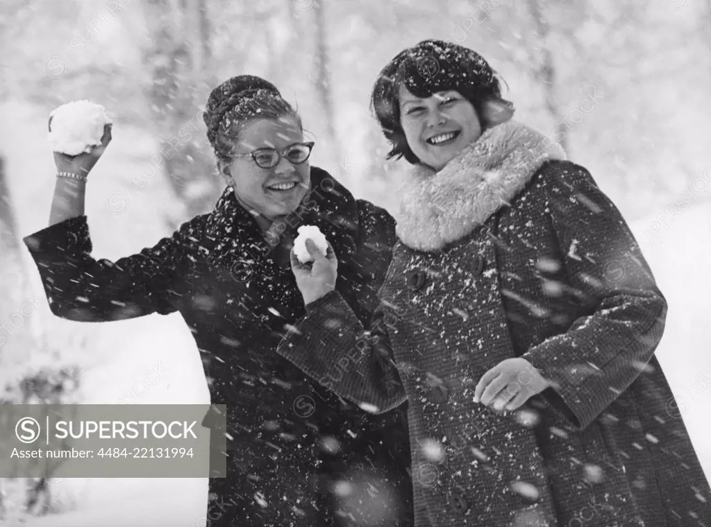 Winter in the 1960s. Two young women is aiming and about to throw snowballs at the photographer. The snow is falling and they are dressed warm with winter coats. Sweden 1964