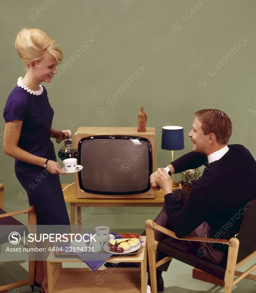 Coffee in the 1960s. A woman is pouring coffee for her husband in front of the tv set. She has her hair in the typical Beehive hairstyle, in which her long hair is piled up on the top of the head and giving some resemblance to the shape of a traditional beehive. The chairs are of the Safari design type. Sweden 1960s