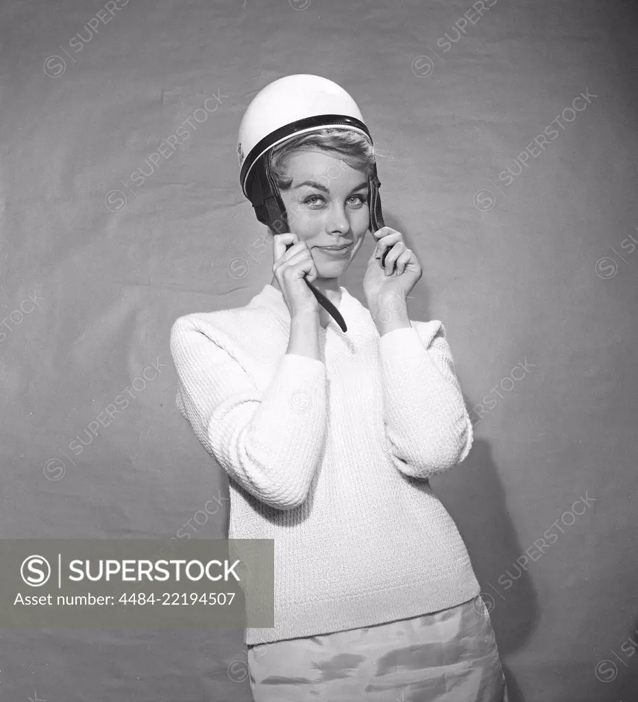 Woman in the 1950s. A young woman is standing dressed in a typical 50s sweater. She is putting on a white moped helmet. Photo Kristoffersson ref BZ8-6. Sweden 1952