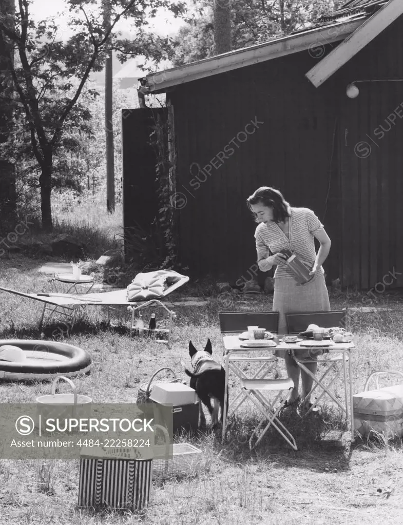 Camping in the 1960s. When camping in the 1960s plastic cups and other camping equipment were sometimes packed and transported in special cases. Pictured a practical such and a woman setting the table for a meal in the nature. The chairs and the table are typical camping folding ones, that could easily be stored and transported, not to take up too much room in the trunk of the car. Every visible object in the picture represents the 1960s decade. Both in material and design. Sweden 1963