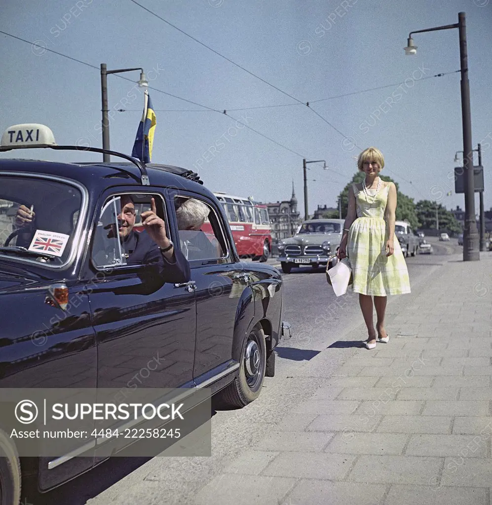 Taxi driver in the 1960s. A taxi driver looks as if he's trying to point out something interesting to the tourist in the back seat. He is however more interested in looking at the blonde young girl on the sidewalk. Sweden 1960 ref BV71-5