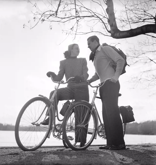 1940s couple on bicycles. A young couple is out on a tour on their bicycles on a sunny spring day. They have practical bags attached to the racks of the bicycles where they could transport the picnic food. Sweden 1947. Photo Kristoffersson AB11-10